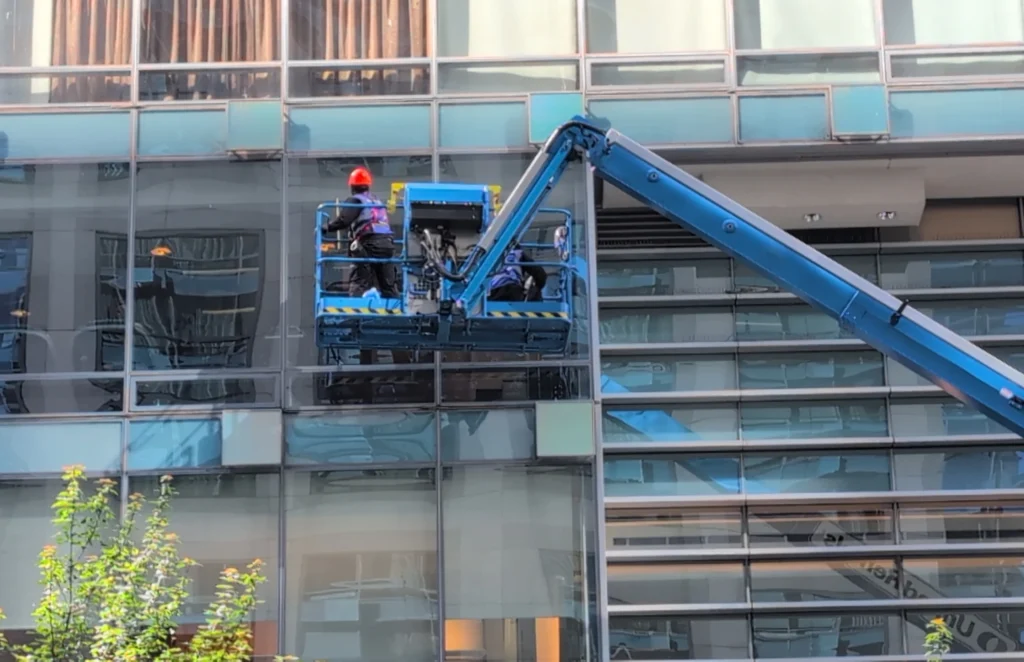 The image shows a window cleaning operation on a glass-fronted building. A blue hydraulic lift (aerial work platform) is raised against the side of a modern building with a glass facade. A worker wearing a bright orange/red safety helmet is standing on the platform of the lift, working on the glass exterior of the building. The hydraulic arm of the lift extends diagonally across the frame. The building features reflective glass panels that show some reflections of the surrounding environment. Some greenery or plants are visible at the bottom left corner of the image. This appears to be a professional window cleaning service in progress, likely related to the commercial cleaning services offered by JF Services.

JF Services | Window Cleaning Vancouver | Window Cleaning | Window Cleaning Canada | Professional Window Cleaning