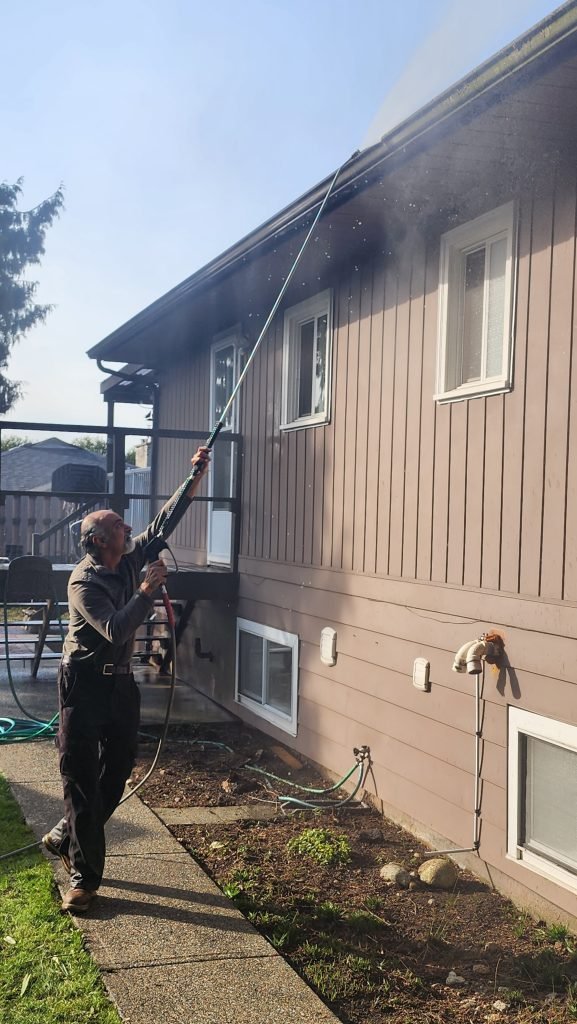 The image shows someone pressure washing or cleaning the exterior of a two-story residential house. The person is using a long extension tool to reach the upper section of the home near the roof line and gutters. Water or cleaning spray can be seen coming from the equipment.

The house has beige/tan siding with white window trim and appears to be a typical suburban home. There's a concrete walkway along the side of the house, with some landscaping beds visible. The sky is clear and blue, suggesting it's a nice day for exterior home maintenance.

This appears to be a professional cleaning service in action, possibly performing pressure washing, exterior washing, or some form of home maintenance. This type of service helps maintain the appearance and longevity of residential properties.

JF Services | Eco Friendly Solutions at JF Services | Eco-Friendly solutions | JF Services | JF Pressure Wash