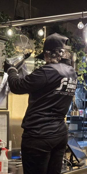 A professional from JF Janitorial Services cleaning a stainless steel kitchen area in Vancouver, BC. The cleaner is wearing a black uniform and using a spray tool to sanitize surfaces. The commercial kitchen features hanging lights, decorative plants, and framed certificates on the wall.